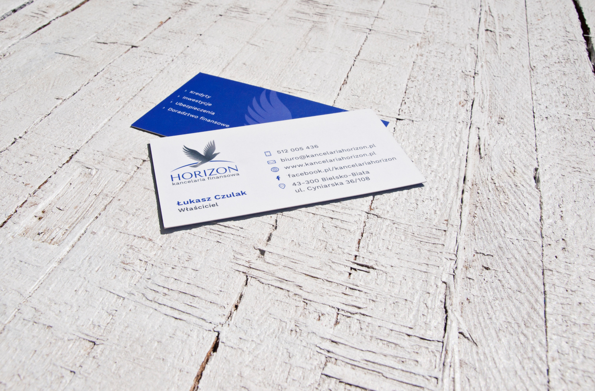 Dwie wizytówki firmy Horizon Kancelaria Finansowa leżące na białym, postarzanym drewnianym tle. Na jednej z nich wypisane usługi: Kredyty, Inwestycje, Ubezpieczenia, Doradztwo finansowe. Na drugiej...