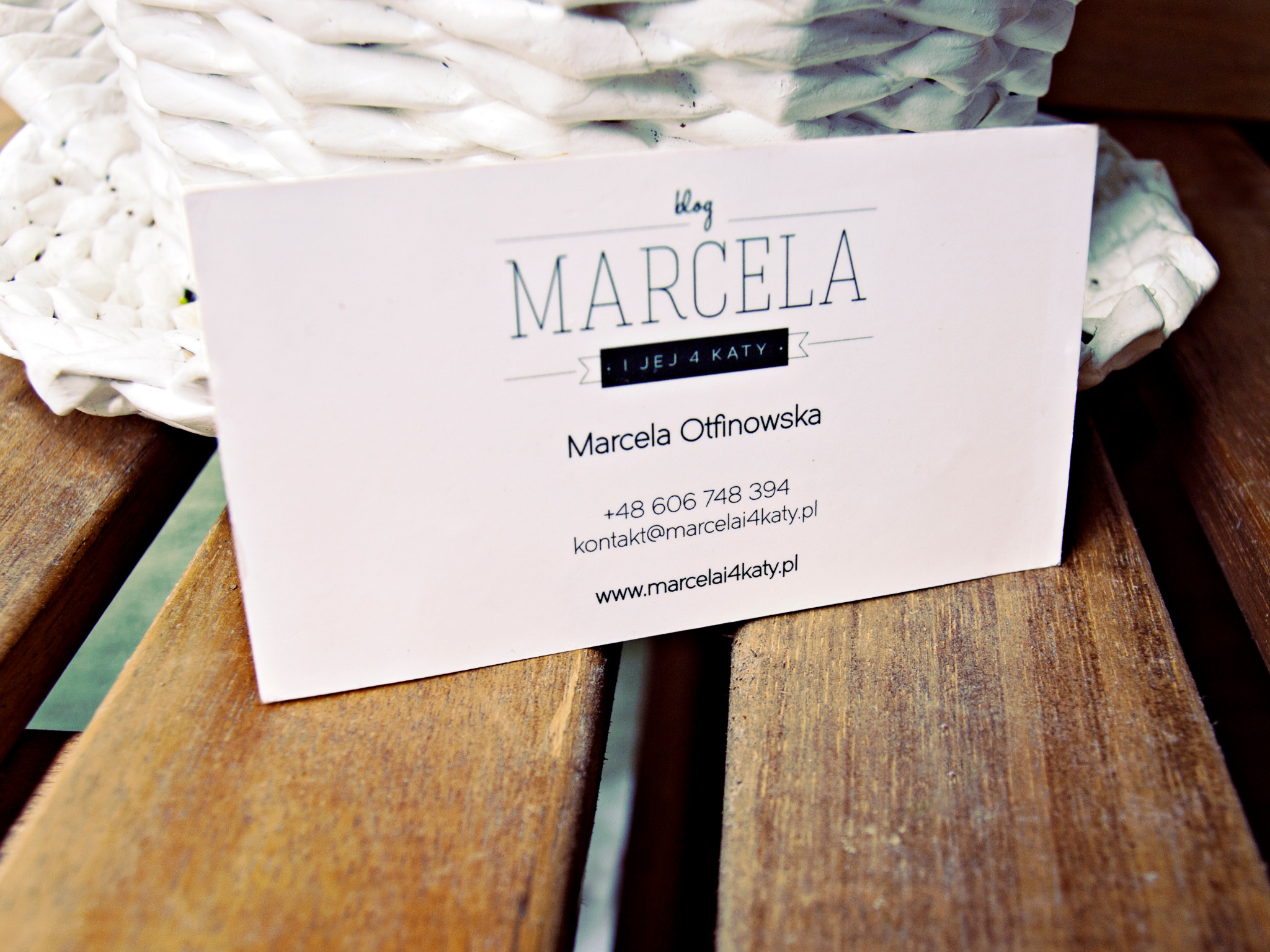 Wizytówka bloga 'Marcela i jej 4 kąty' leży na drewnianej powierzchni, w tle biały, pleciony kosz.