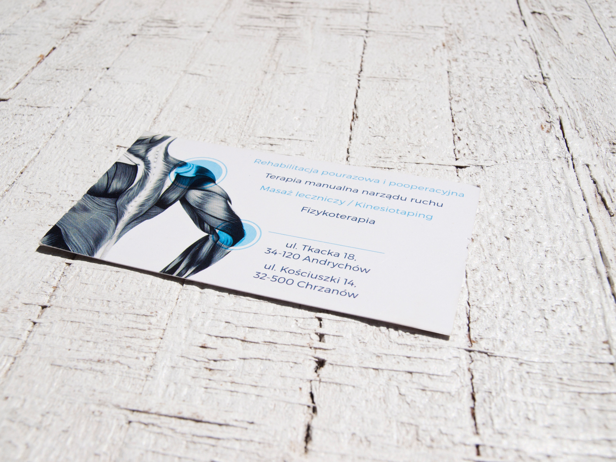 Wizytówka z ilustracją mięśni i informacjami o rehabilitacji pourazowej i pooperacyjnej, terapii manualnej, masażu leczniczym, kinesiotapingu i fizykoterapii, leżąca na białym drewnianym tle.