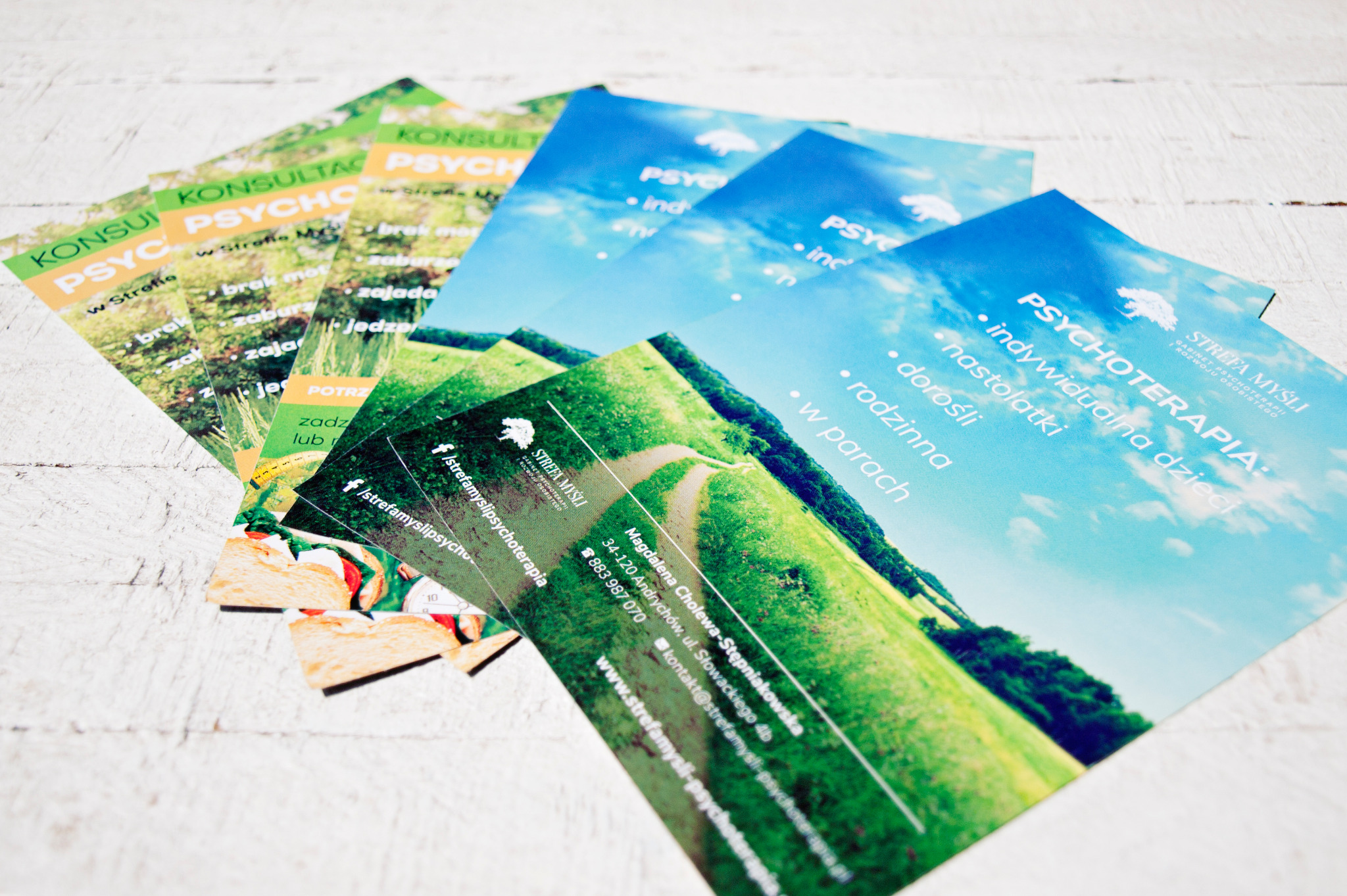 Stos ulotek i wizytówek gabinetu psychoterapii 'Strefa Myśli' z motywami krajobrazowymi i informacjami kontaktowymi, ułożonych na białym, drewnianym tle.