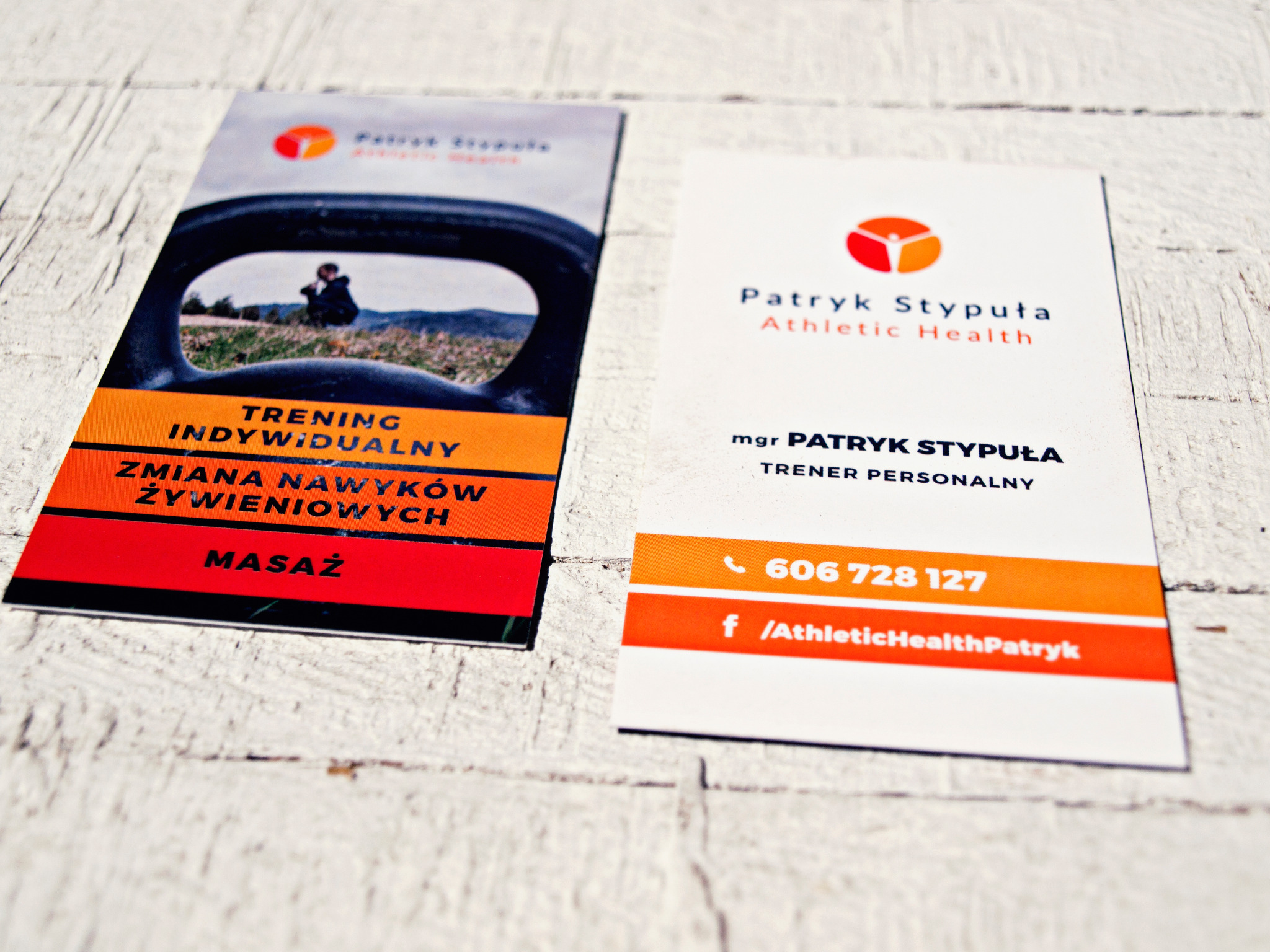 Dwie wizytówki na białym, drewnianym tle. Jedna wizytówka z grafiką osoby ćwiczącej z kettlebell, druga z danymi kontaktowymi trenera personalnego Patryka Stypuły.