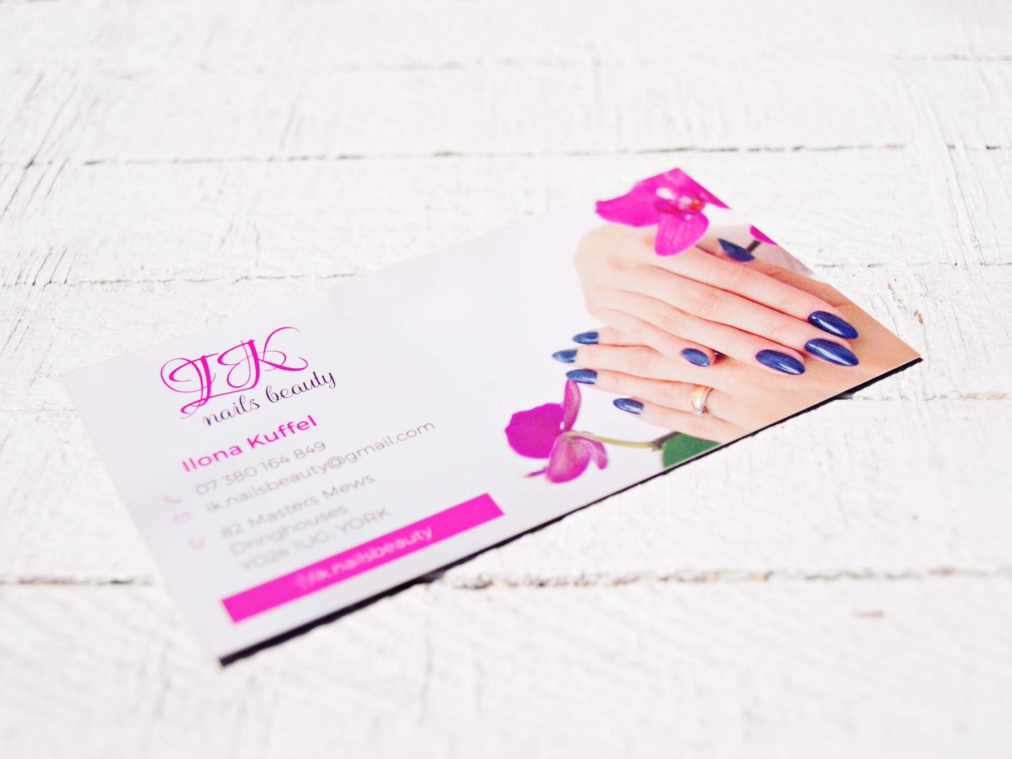 Wizytówka firmy kosmetycznej z grafiką dłoni z ciemnoniebieskim manicure i różowym kwiatem orchidei, leżąca na białym drewnianym tle.