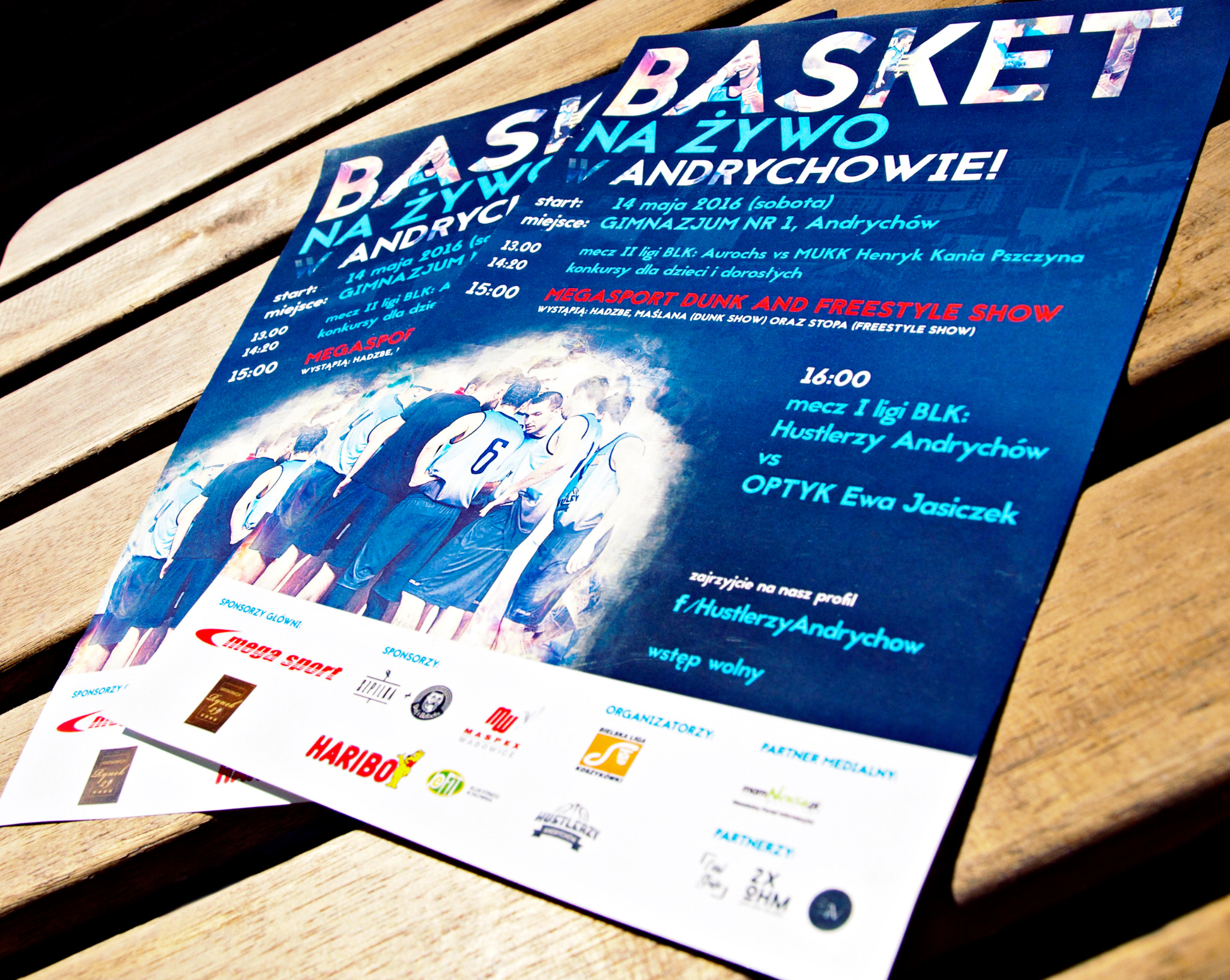 Dwa plakaty z informacjami o wydarzeniu sportowym BASKET NA ŻYWO W ANDRYCHOWIE! leżą na drewnianym stole. Plakat zawiera szczegóły dotyczące meczów, konkursów i sponsorów.