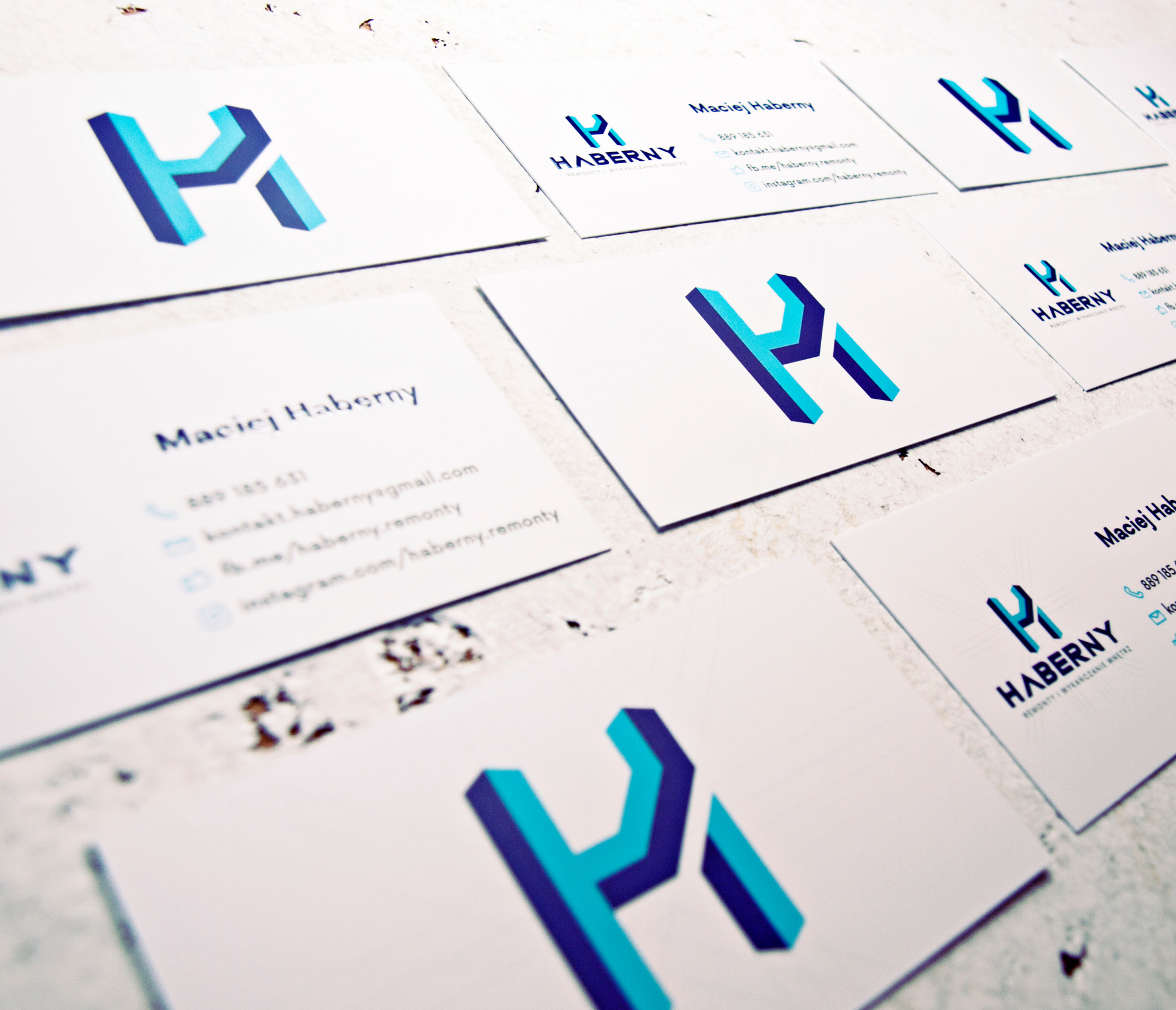 Układ kilku wizytówek firmy Haberny z trójwymiarowym, geometrycznym logo w odcieniach niebieskiego, ułożonych na teksturowanej powierzchni.