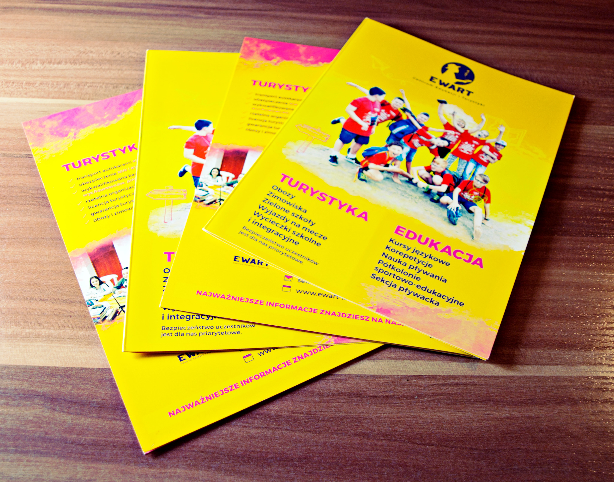 Stos żółtych ulotek z informacjami o obozach i wycieczkach szkolnych firmy z Andrychowa, ułożonych na drewnianym blacie pod lekkim kątem.
