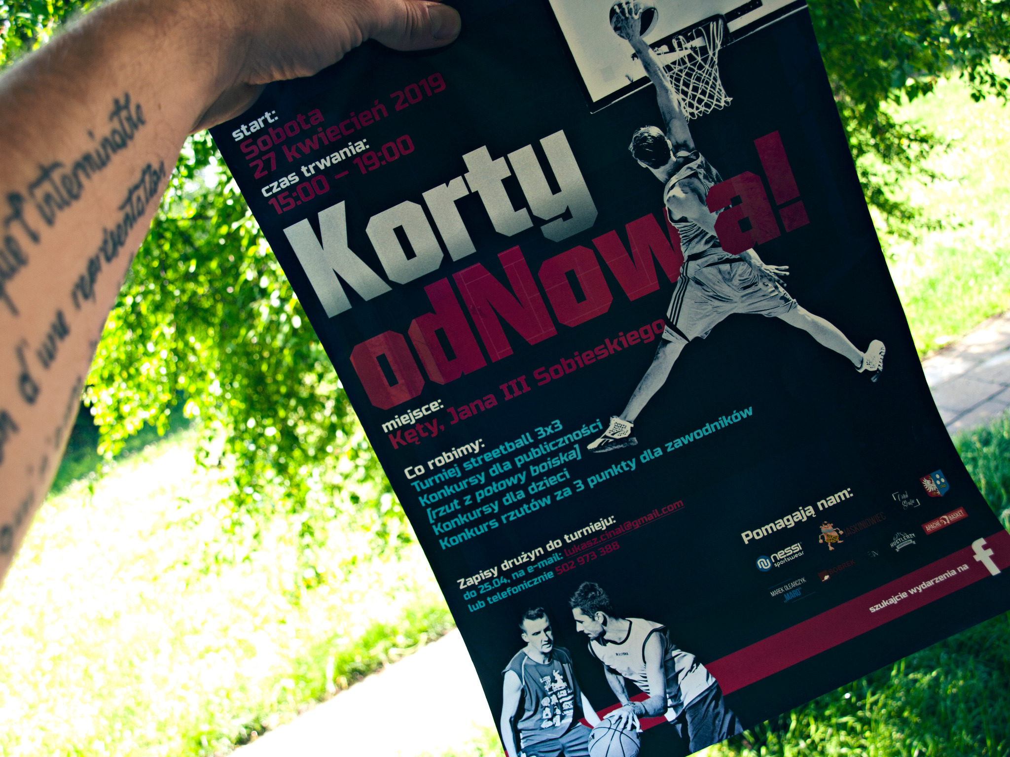 Ręka z tatuażem trzyma plakat reklamujący turniej streetball 3x3 'Korty odNowa!' z datą 27 kwietnia 2019, miejscem Kęty i informacjami o konkursach dla publiczności i dzieci.
