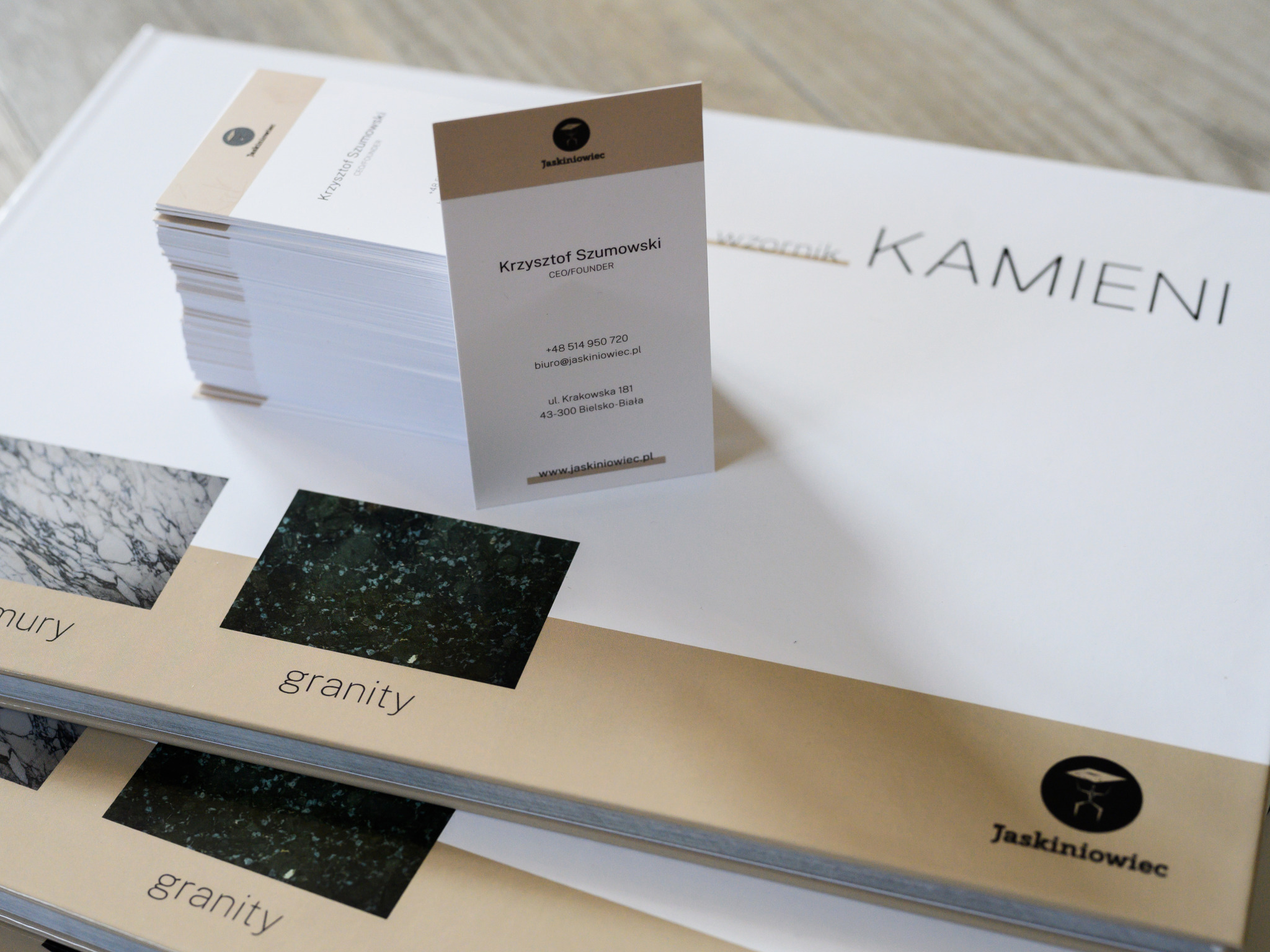 Stos wizytówek firmy Jaskiniowiec na tle wzornika kamieni naturalnych z próbkami granitu i marmuru.