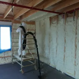 Pracownik w kombinezonie ochronnym na rusztowaniu aplikuje piankę izolacyjną na ściany wewnętrzne budynku w trakcie budowy, widoczne drewniane elementy konstrukcyjne i czerwone rury pod sufitem.