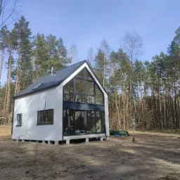 Nowoczesny, biały dom z dużymi oknami na fundamencie punktowym, otoczony lasem sosnowym w słoneczny dzień.