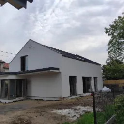 Surowy stan domu jednorodzinnego z białym tynkiem, czarnym dachem, dużymi oknami i widocznymi śladami prac budowlanych na terenie posesji.