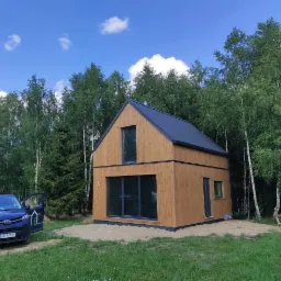 Nowoczesny, dwukondygnacyjny dom szkieletowy z drewnianą elewacją i ciemnym dachem, otoczony drzewami, z błękitnym samochodem z logo firmy w tle.