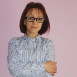 Portret kobiety w okularach, z rękami skrzyżowanymi na tle różowego tła. Ubrana w koszulę w paski z drobnymi wzorami. Widoczne kolczyki i pierścionek.