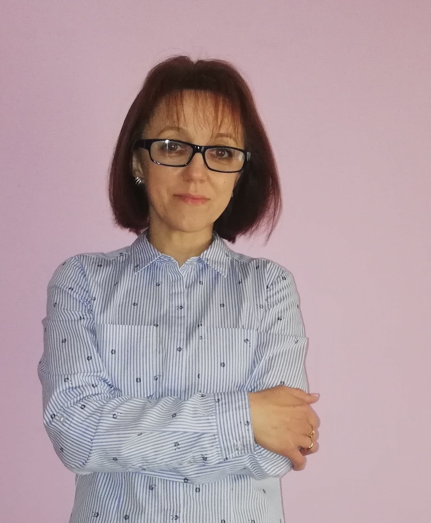 Portret kobiety w okularach, z rękami skrzyżowanymi na tle różowego tła. Ubrana w koszulę w paski z drobnymi wzorami. Widoczne kolczyki i pierścionek.