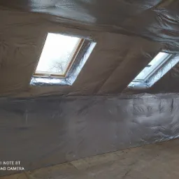 Poddasze z oknami dachowymi w trakcie ocieplania folią termoizolacyjną, widoczne szwy i niedociągnięcia w montażu folii. W tle widoczne drzewo za oknem.