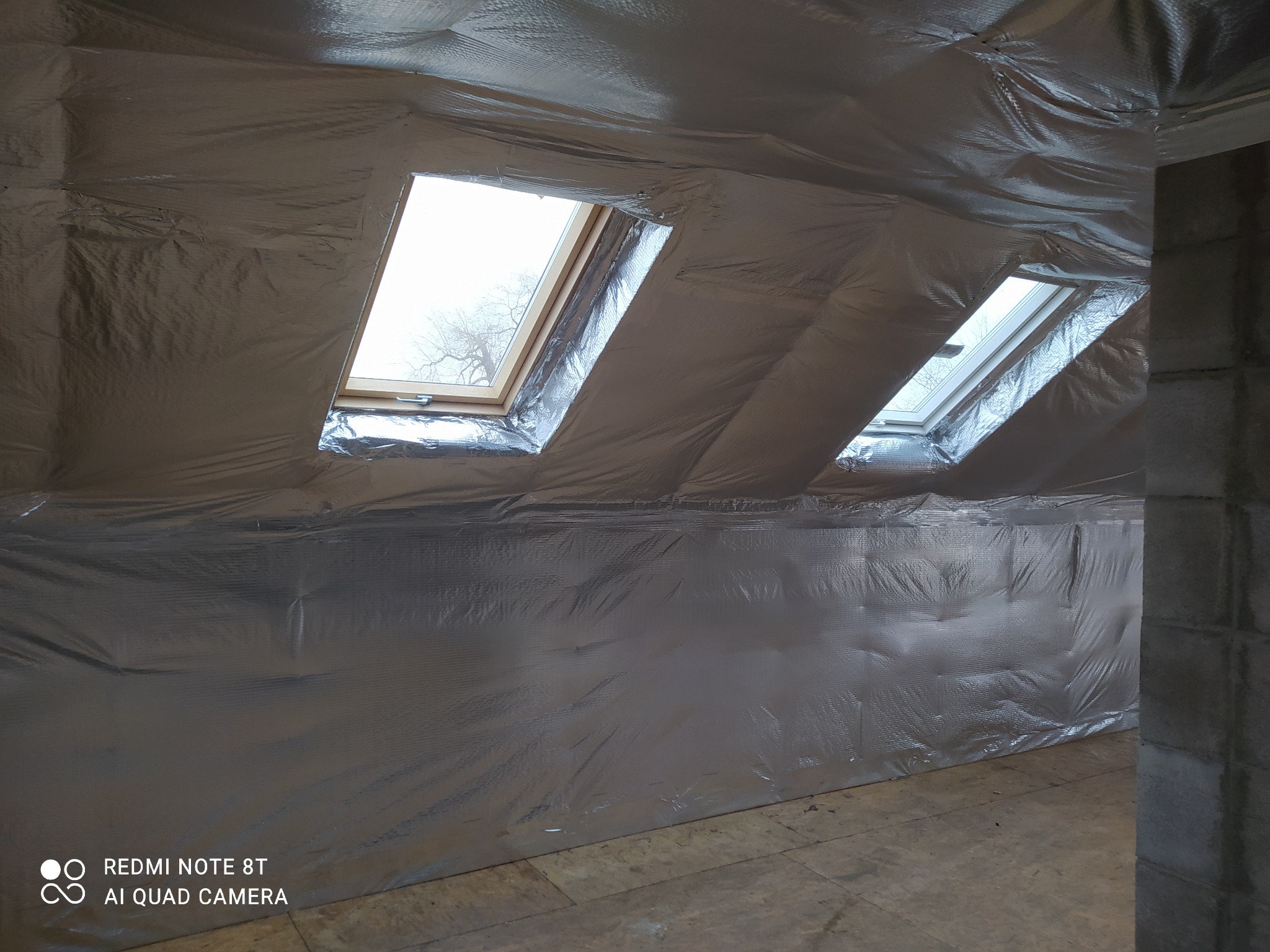 Poddasze z oknami dachowymi w trakcie ocieplania folią termoizolacyjną, widoczne szwy i niedociągnięcia w montażu folii. W tle widoczne drzewo za oknem.