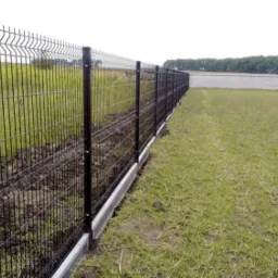 Długi, czarny panelowy płot z siatki, osadzony na betonowej podmurówce, oddziela uprawne pole od trawnika. W tle widoczny mur i drzewa.