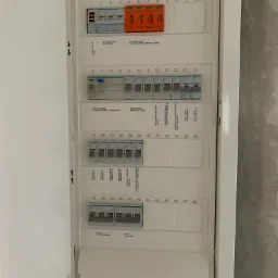 Otwarta skrzynka elektryczna z bezpiecznikami i wyłącznikami różnicowoprądowymi, widoczne opisy obwodów, instalacja w trakcie prac wykończeniowych.