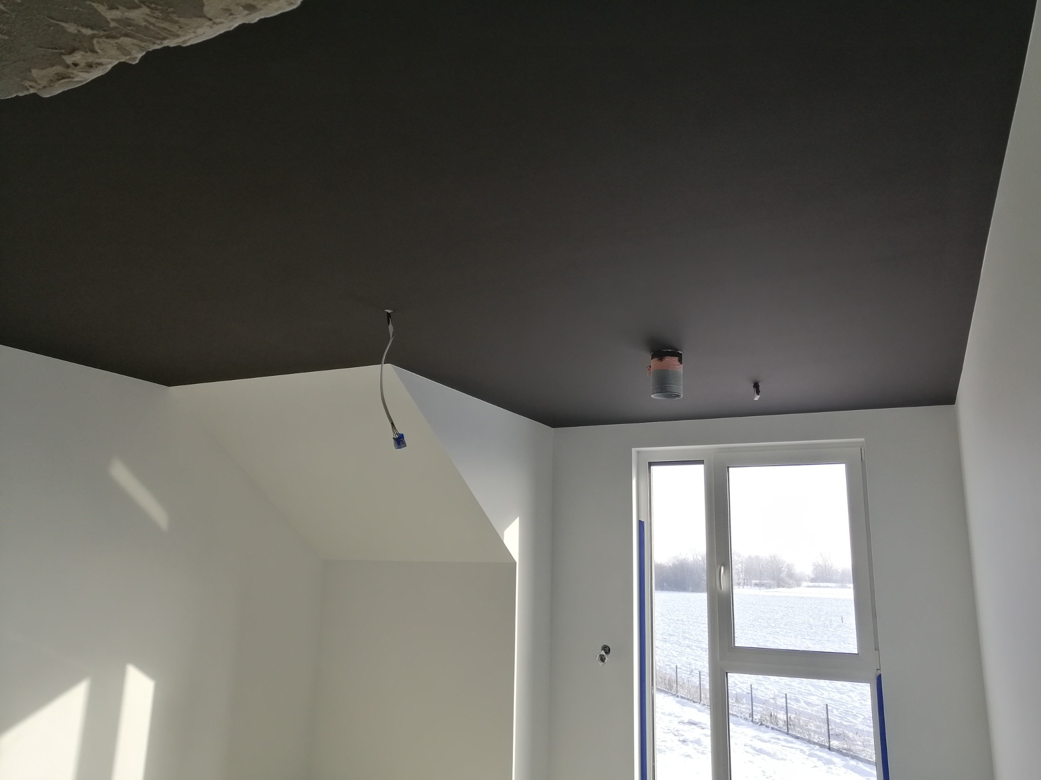 Pokój po remoncie z pomalowanym na ciemno sufitem, białe ściany, widoczne okno z zimowym krajobrazem za szybą, wystające kable i otwory po lampach na suficie.