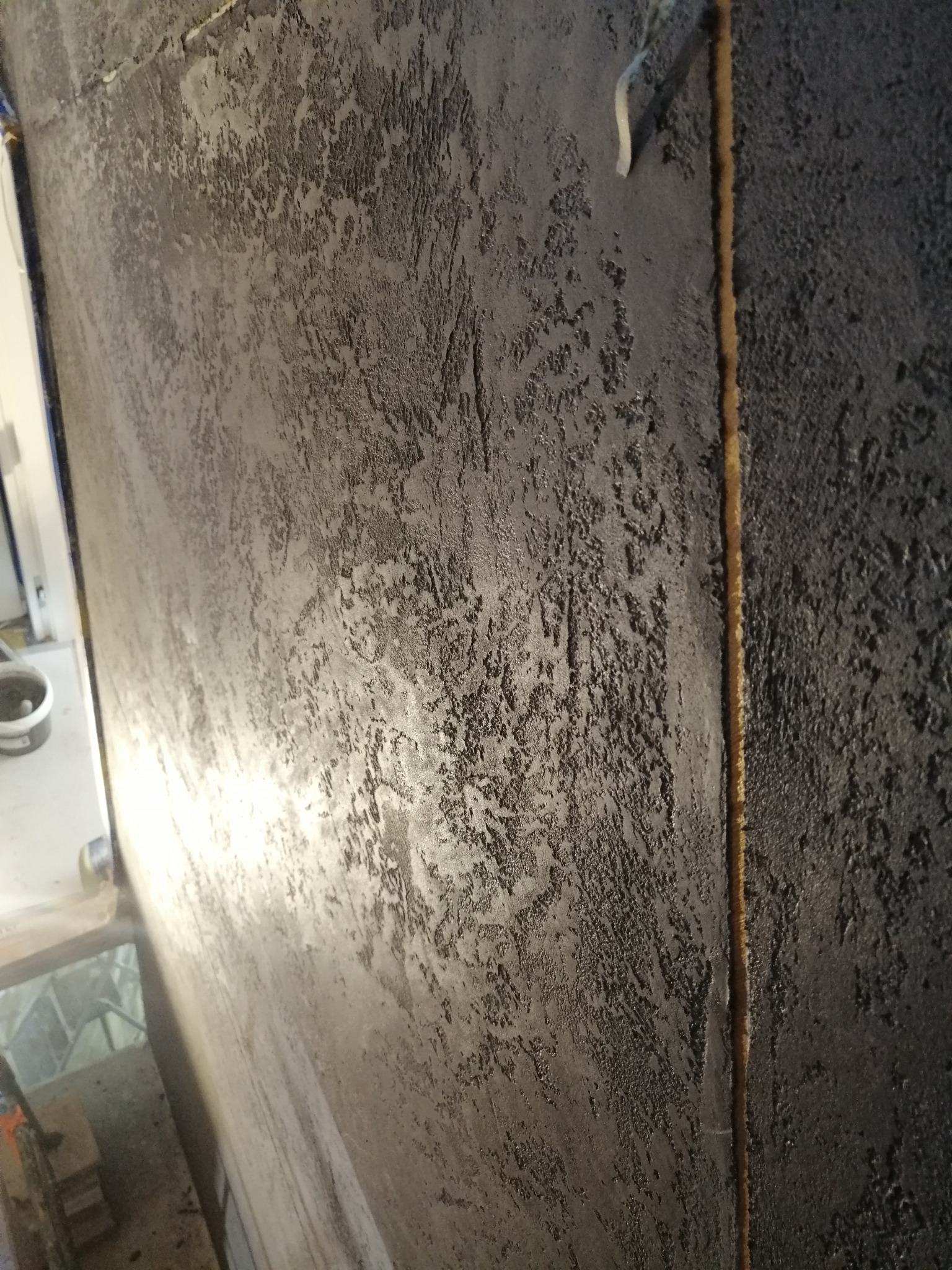 Ściana z ciemną, strukturalną powłoką dekoracyjną, z wyraźną pionową linią łączenia dwóch powierzchni, widoczne niedoskonałości faktury i odbicie światła.