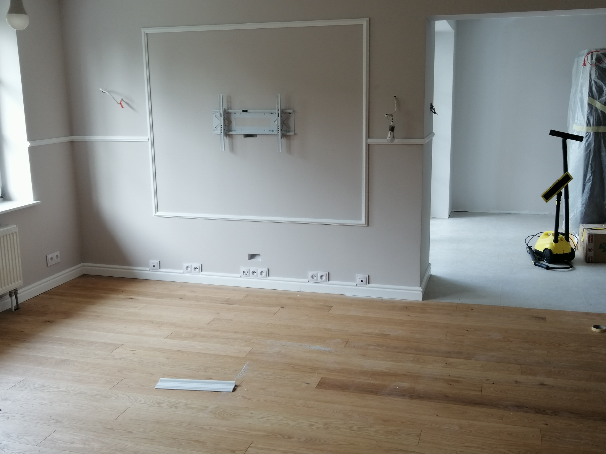 Pokój po remoncie: świeżo położona podłoga z paneli, pomalowane ściany z dekoracyjną sztukaterią i zamontowanym uchwytem na telewizor, widoczne gniazdka elektryczne przy podłodze, w tle odkurzacz...