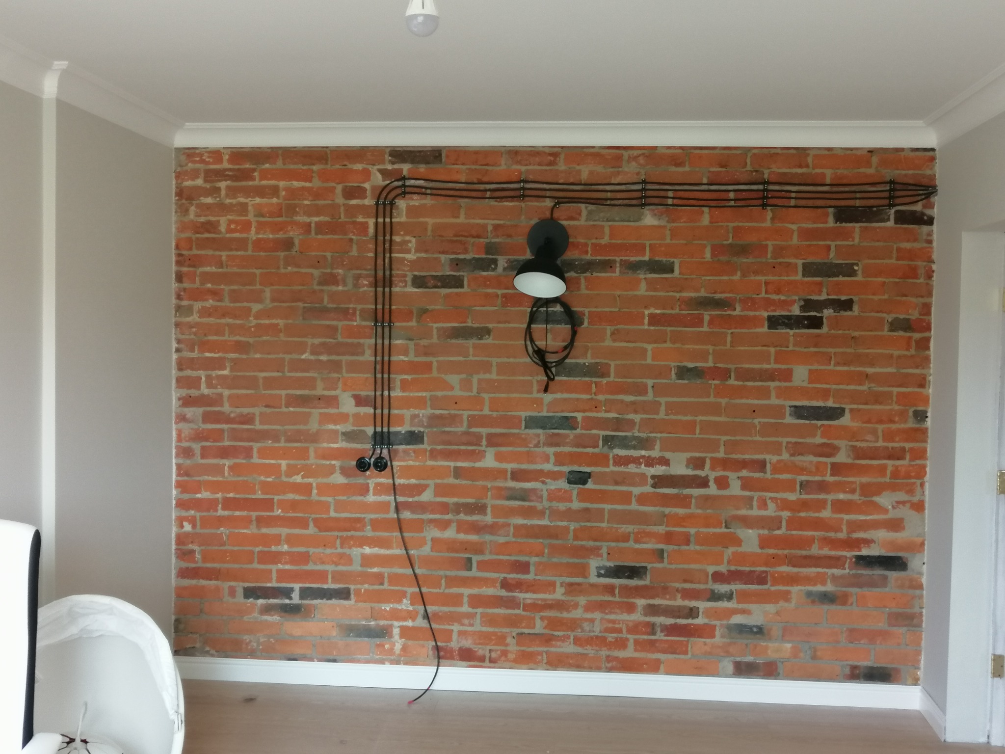 Ceglana ściana z widocznymi przewodami elektrycznymi, czarną lampą ścienną i gniazdkami, surowy industrialny styl, białe listwy przypodłogowe i sufitowe
