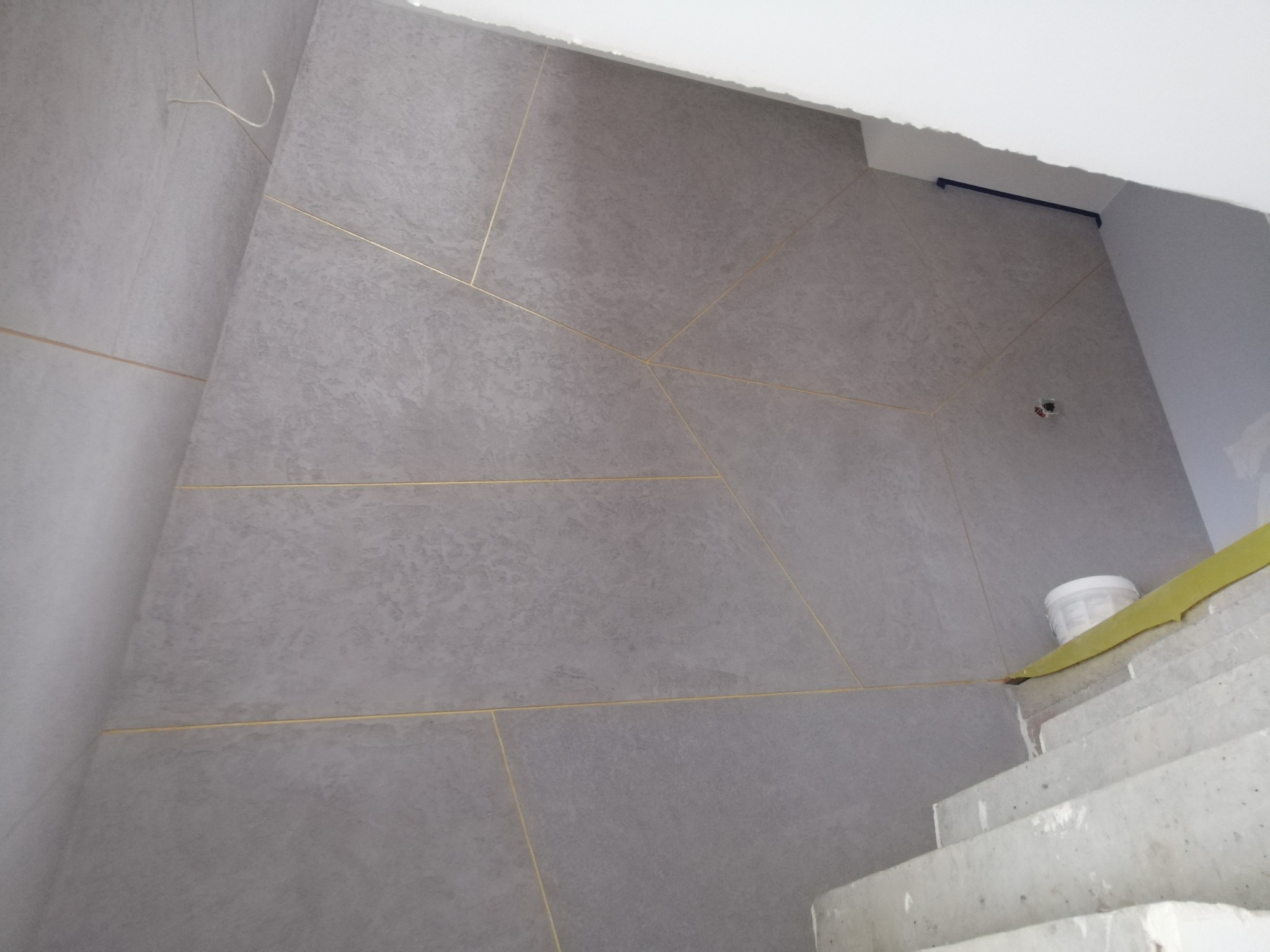 Ściana wykończona szarymi płytami z delikatną fakturą, spoiny podkreślone złotymi listwami, widoczne schody i wiadro z farbą w narożniku, częściowo zakryte żółtą taśmą malarską.