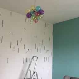 Krzysztof Krasowski - Pokój w trakcie odświeżania: ściana z motywem leśnym i drabina aluminiowa, obok ściana pomalowana na zielono, pod sufitem lampa w kształcie balonu z kolorowych kul.