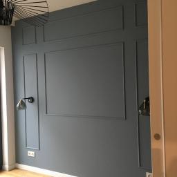 Krzysztof Krasowski - Szara ściana z dekoracyjnymi listwami w stylu angielskim, drewniana podłoga, nowoczesna lampa sufitowa i kinkiety w kolorze grafitowym.