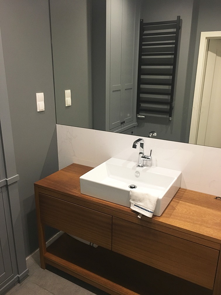 Nowoczesna łazienka z kwadratową umywalką nablatową na drewnianej szafce, widoczne szare ściany, lustro z odbiciem szafki i grzejnika drabinkowego.