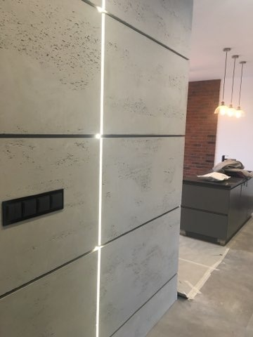 Ściana wykończona płytami o strukturze betonu z wbudowanym oświetleniem LED, czarny włącznik światła po lewej stronie, widok na wnętrze z ceglaną ścianą w tle.