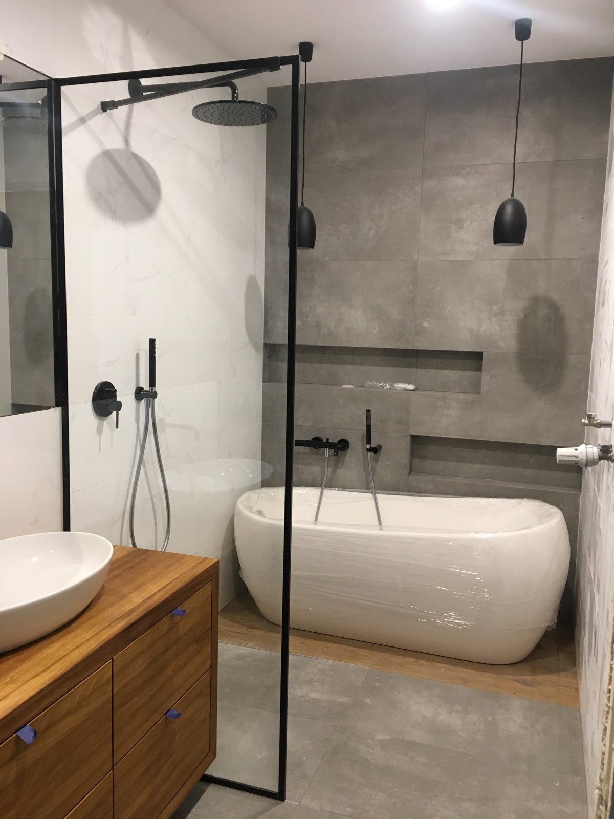 Nowoczesna łazienka w szaro-białej kolorystyce z wolnostojącą wanną owiniętą folią ochronną, czarną armaturą i kabiną prysznicową typu walk-in z czarnymi profilami, drewniana szafka pod umywalkę...