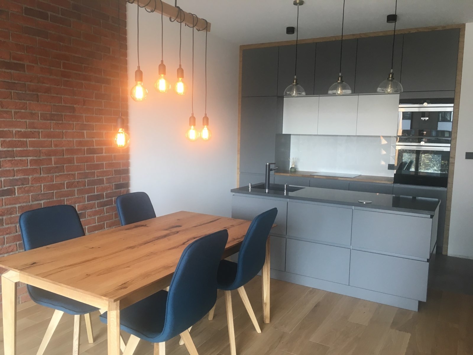 Nowoczesna kuchnia z szarą zabudową, drewnianym stołem i ceglaną ścianą, oświetlona wiszącymi lampami z żarówkami Edisona.