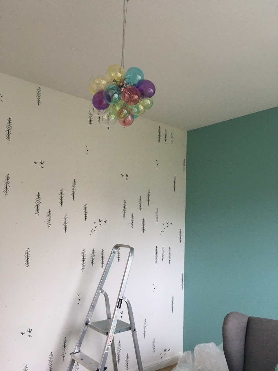 Pokój w trakcie odświeżania: ściana z motywem leśnym i drabina aluminiowa, obok ściana pomalowana na zielono, pod sufitem lampa w kształcie balonu z kolorowych kul.