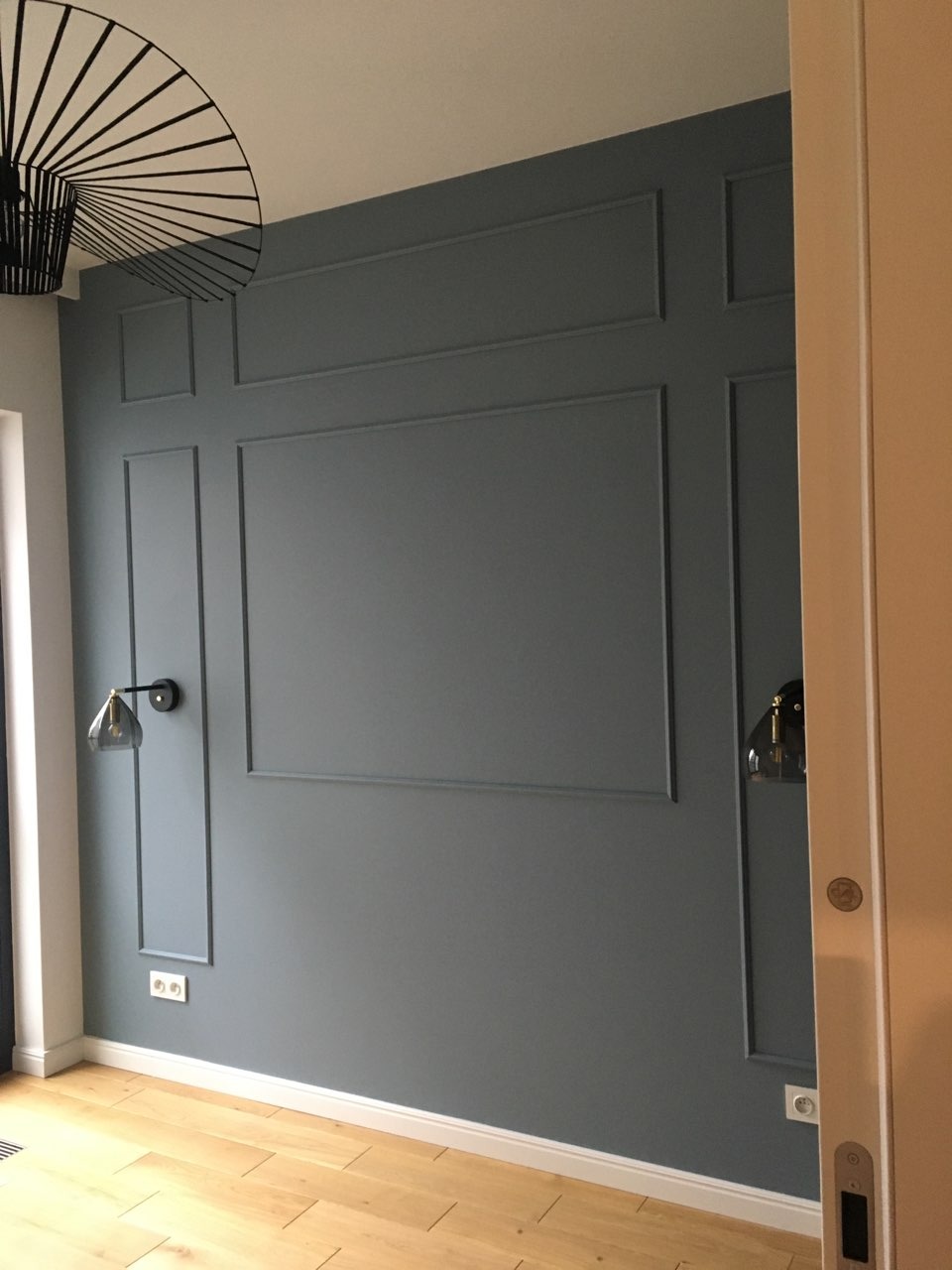 Szara ściana z dekoracyjnymi listwami w stylu angielskim, drewniana podłoga, nowoczesna lampa sufitowa i kinkiety w kolorze grafitowym.