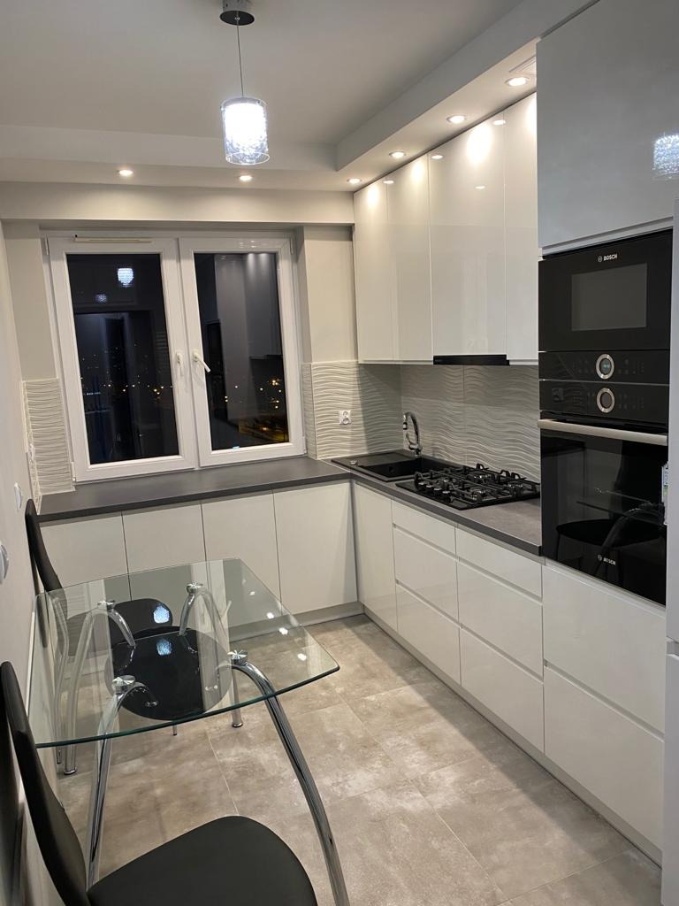 Nowoczesna, jasna kuchnia z białymi, lakierowanymi szafkami, szarym blatem i szklanym stołem z czarnymi krzesłami, oświetlona wiszącą lampą i punktowymi światłami LED.