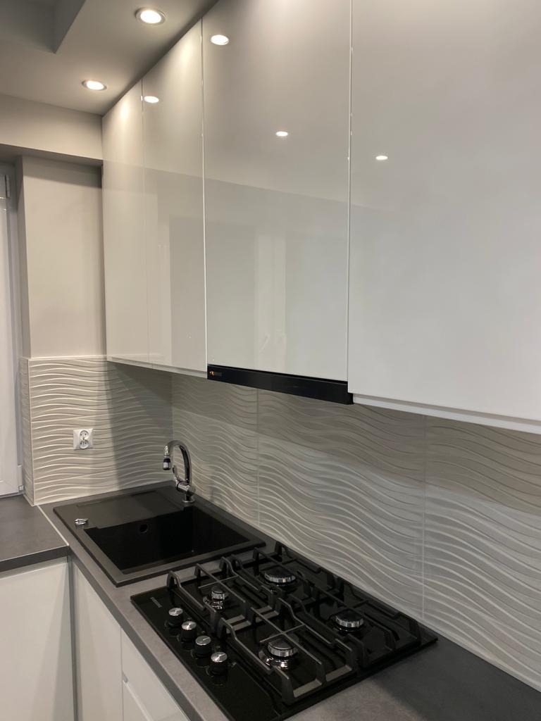 Nowoczesna kuchnia z białymi, lakierowanymi szafkami wiszącymi, czarnym zlewem i płytą gazową, oraz falistymi płytkami ściennymi