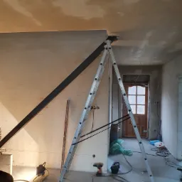 Wnętrze w trakcie remontu, widoczna metalowa belka podpierająca sufit, drabina aluminiowa, narzędzia budowlane i nieotynkowane ściany.