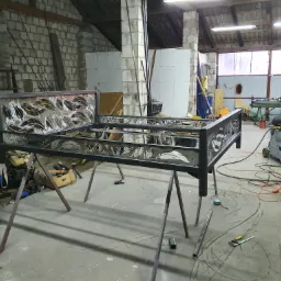 Metalowa konstrukcja łóżka z dekoracyjnymi wycięciami w kształcie liści, w trakcie spawania w warsztacie, widoczne narzędzia i spawarka, surowy mur z cegieł w tle.