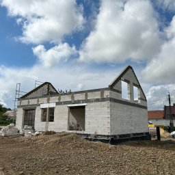 Inżynieria Budowlana Jędrzej Radziewicz - Surowy stan domu z białych bloczków, widoczne otwory okienne i zarys dachu na tle błękitnego nieba z chmurami, stos bloczków na ziemi, teren budowy.