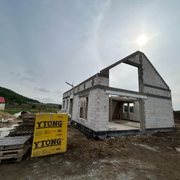 Inżynieria Budowlana Jędrzej Radziewicz - Surowy stan domu murowanego z bloczków Ytong, widoczne palety z materiałem budowlanym, w tle krajobraz wiejski i pochmurne niebo.