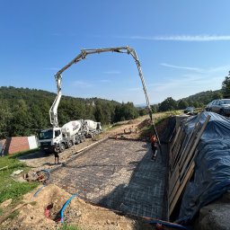 Inżynieria Budowlana Jędrzej Radziewicz - Wylewanie betonu do szalunku zbrojonego siatką stalową na budowie fundamentów domu, z użyciem pompy do betonu z długim ramieniem; w tle widoczny mikser betonu, pracownicy w kaskach, krajobraz...