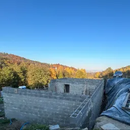 Wznoszone ściany budynku z bloczków betonowych na fundamencie, z widocznym zbrojeniem, w tle jesienny krajobraz zalesionych wzgórz i błękitne niebo.