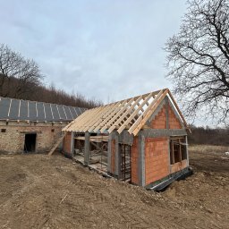 Inżynieria Budowlana Jędrzej Radziewicz - Budowa domu z czerwonej cegły, widoczne ściany z otworami okiennymi, drewniana konstrukcja dachu w trakcie montażu, obok budynek gospodarczy z ciemnym dachem, teren budowy z ziemią i śladami opon...