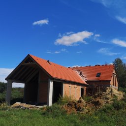 Inżynieria Budowlana Jędrzej Radziewicz - Stan surowy otwarty domu z czerwonym dachem, widoczne pustaki i betonowe słupy, na dachu pracownik układa dachówkę, w tle błękitne niebo z chmurami.