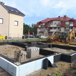 Inżynieria Budowlana Jędrzej Radziewicz - Budowa fundamentów domu w Pieszycach, widoczne izolowane ściany fundamentowe, wystające pręty zbrojeniowe i koparka w tle.