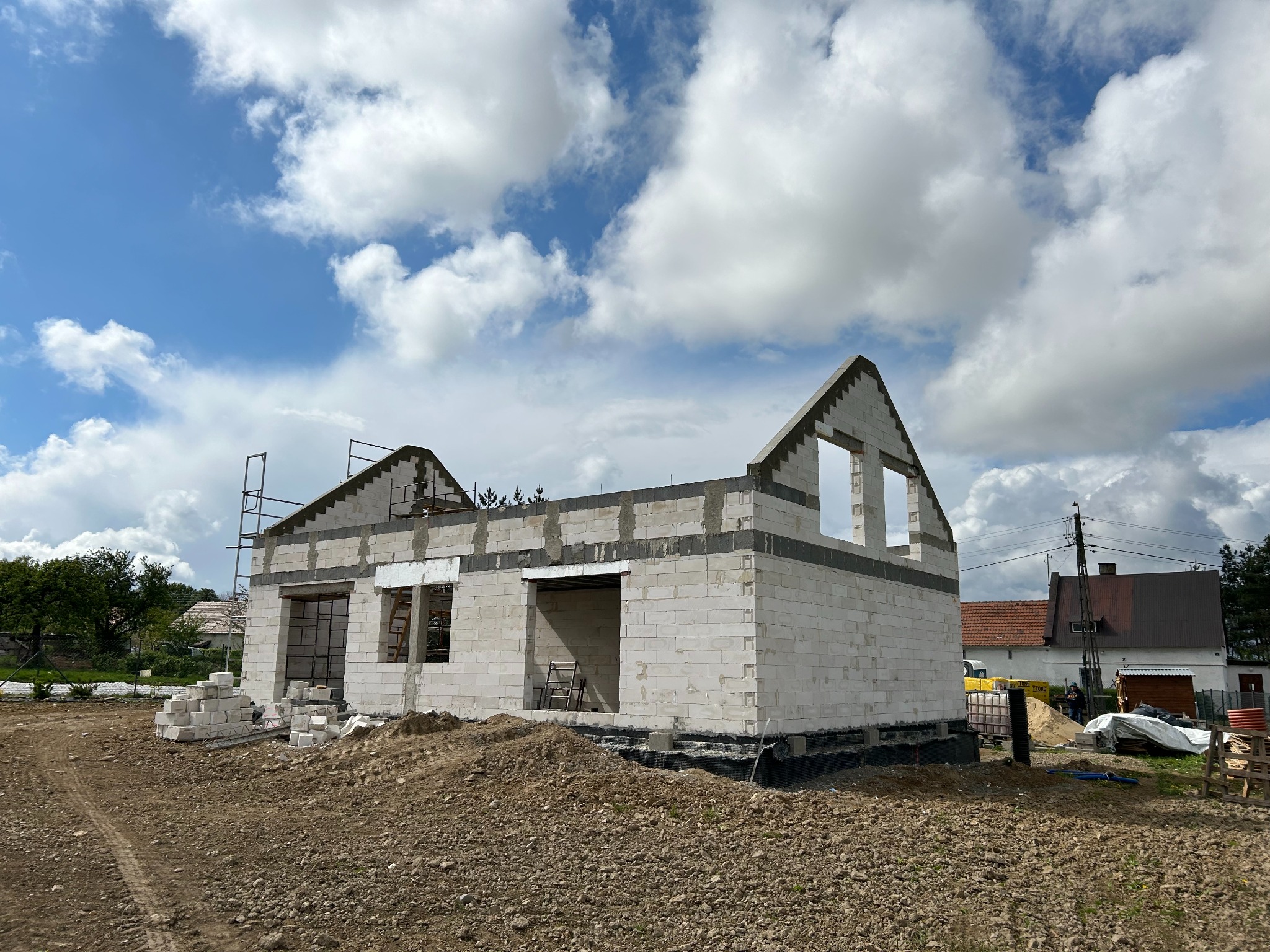 Surowy stan domu z białych bloczków, widoczne otwory okienne i zarys dachu na tle błękitnego nieba z chmurami, stos bloczków na ziemi, teren budowy.