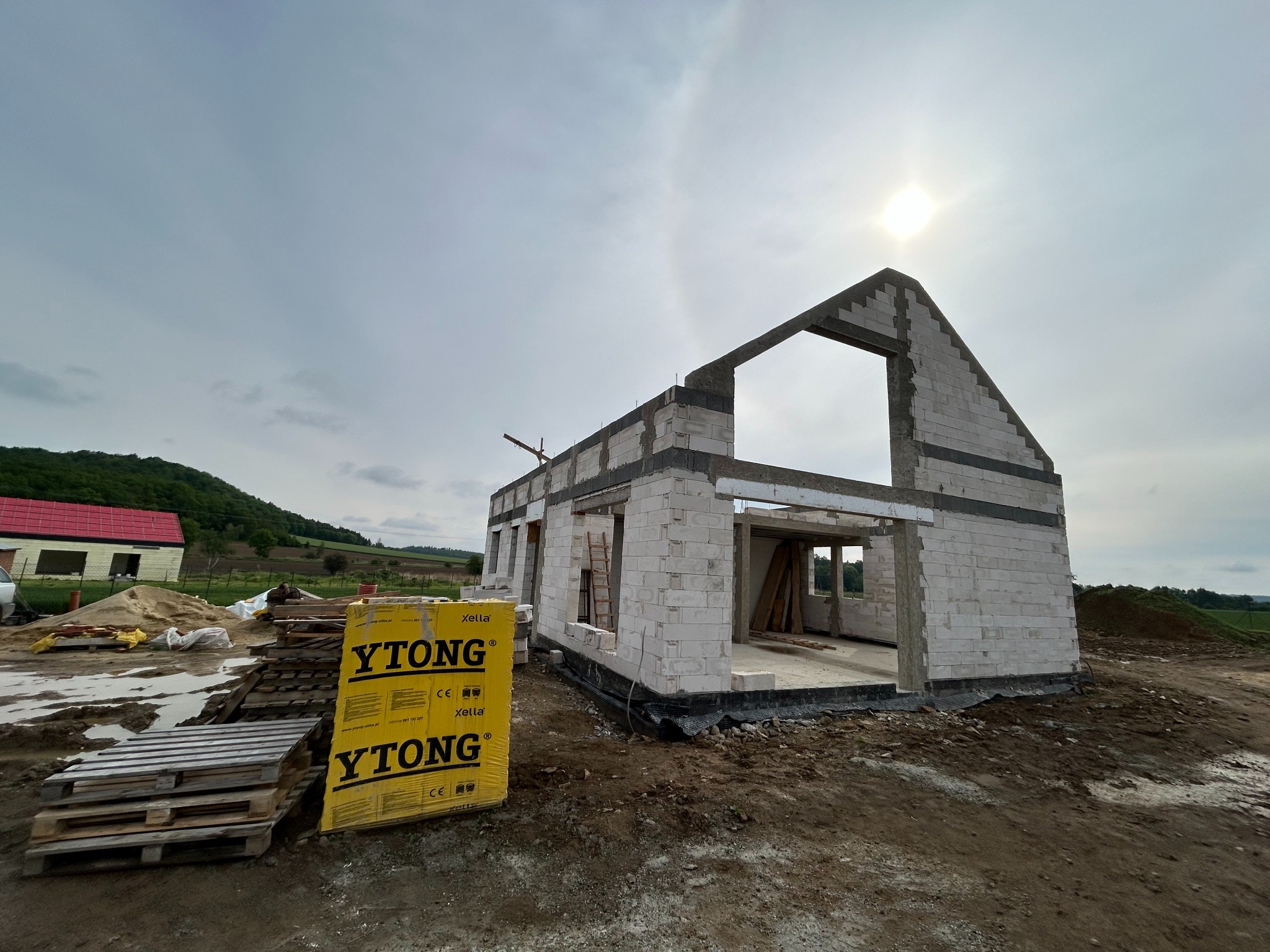 Surowy stan domu murowanego z bloczków Ytong, widoczne palety z materiałem budowlanym, w tle krajobraz wiejski i pochmurne niebo.