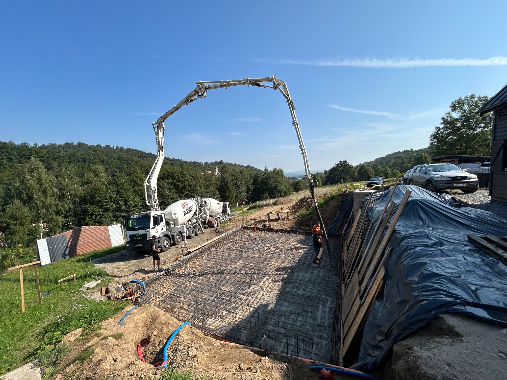 Wylewanie betonu do szalunku zbrojonego siatką stalową na budowie fundamentów domu, z użyciem pompy do betonu z długim ramieniem; w tle widoczny mikser betonu, pracownicy w kaskach, krajobraz...