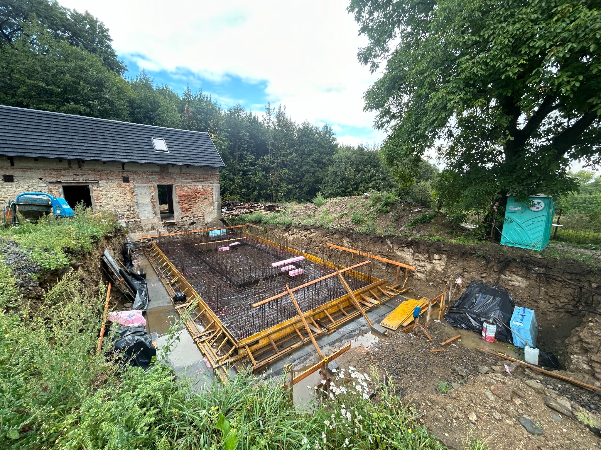 Zbrojenie fundamentu w wykopie z drewnianym szalunkiem, widok na stary budynek gospodarczy z cegły i zielone otoczenie.