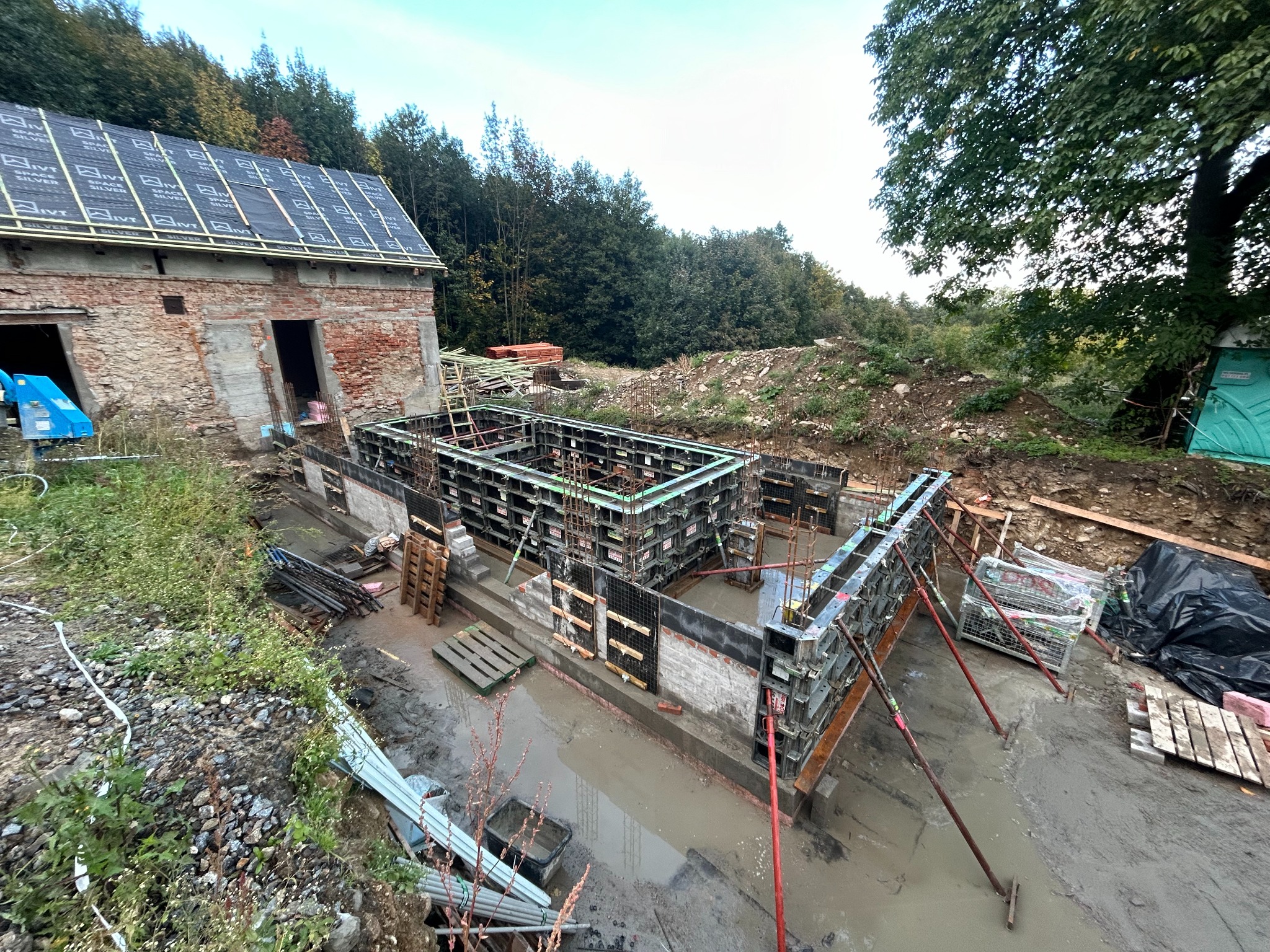 Szare szalunki fundamentowe ustawione na mokrym placu budowy z widoczną starą ceglaną zabudową w tle, częściowo zdemontowane deskowanie i pozostałości po wykopie.
